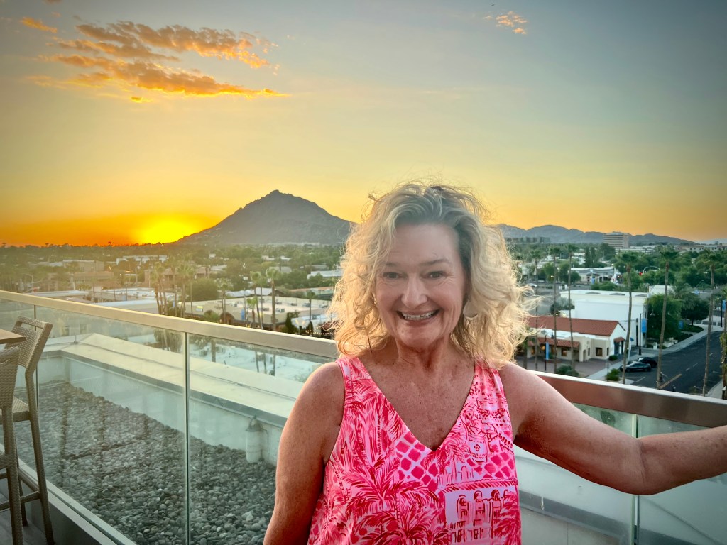 A smiling person with curly hair stands on a balcony at sunset, wearing a pink patterned top, with a mountainous landscape and city in the background.