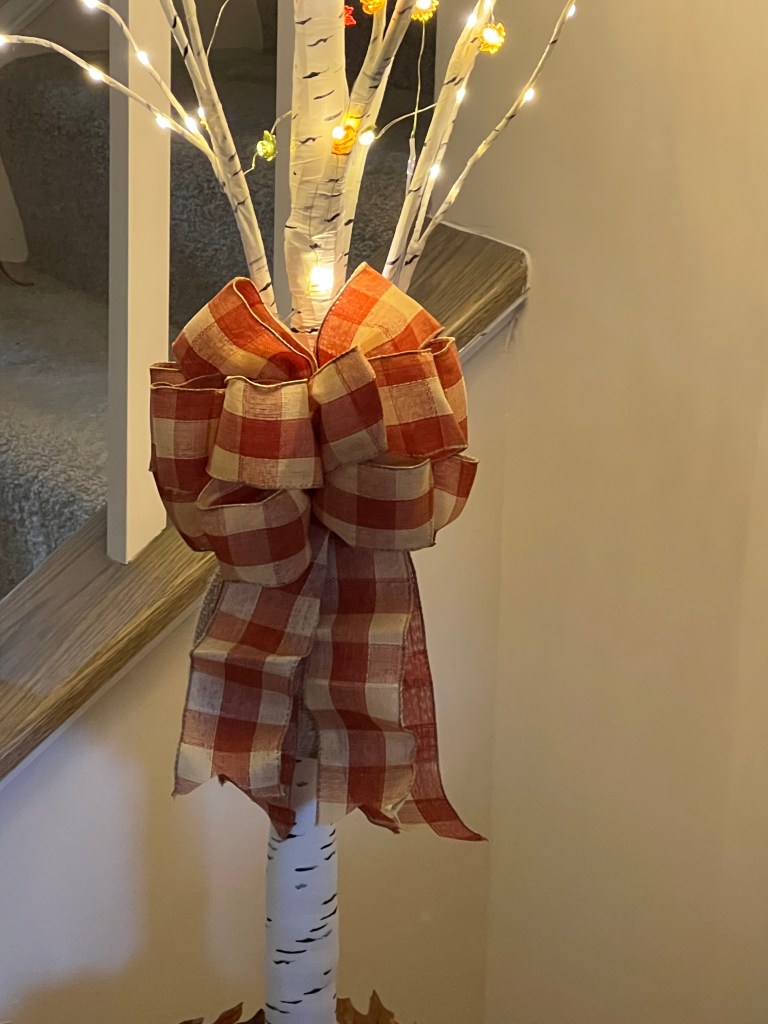 A decorative birch tree adorned with warm-colored fairy lights and topped with a large red and white checkered bow, set against a softly lit staircase.