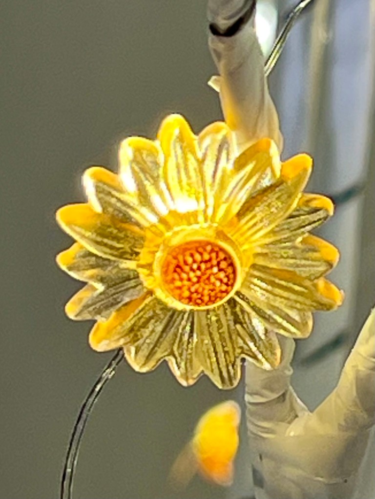 A close-up image of a yellow daisy decoration, featuring textured petals and a bright orange center, surrounded by soft lighting.