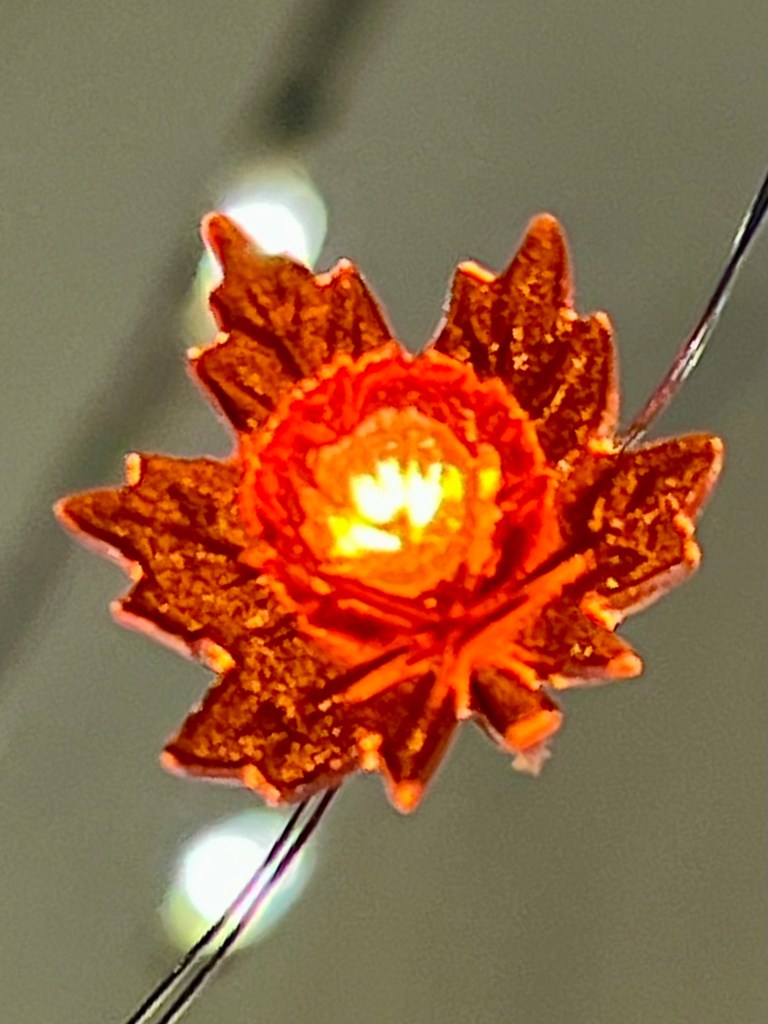 A close-up of a decorative autumn leaf light, glowing with a warm orange hue against a soft blurred background.