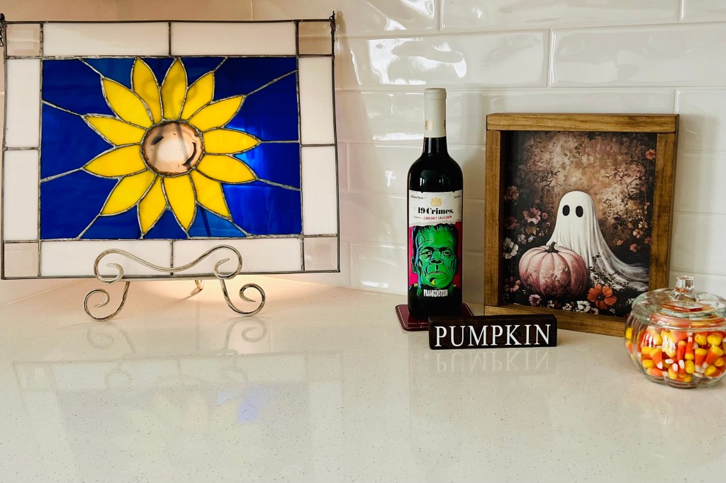 A kitchen countertop decorated for fall, featuring a stained glass sunflower, a Halloween-themed wine bottle, a framed ghost artwork with a pumpkin, a sign that reads 'PUMPKIN', and a jar filled with candy corn.