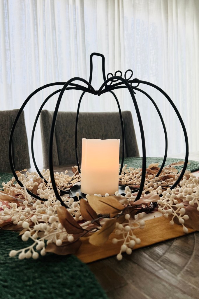 A decorative black wire pumpkin centerpiece surrounding a lit candle, placed on a table with dried leaves and white berries, evoking a cozy autumn atmosphere.