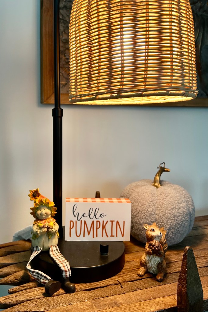 A cozy autumn-themed tabletop display featuring a lamp with a woven shade, a decorative sign that reads 'hello PUMPKIN,' a cute scarecrow figure, a plush gray pumpkin, and a small squirrel figurine holding an acorn.