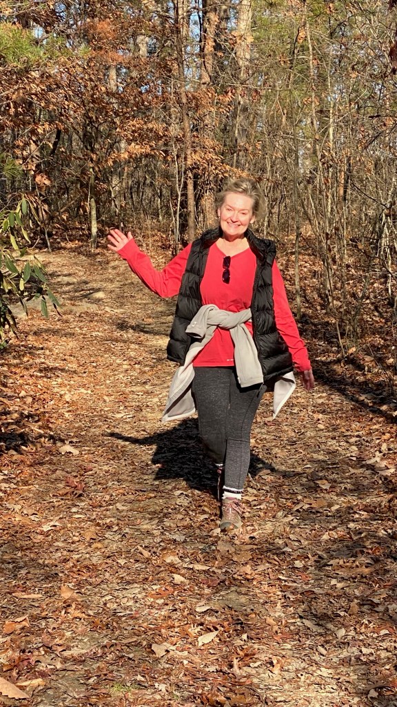 A person walking along a leaf-covered path in the woods during autumn, wearing a red top, black vest, and gray leggings, with a joyful expression.