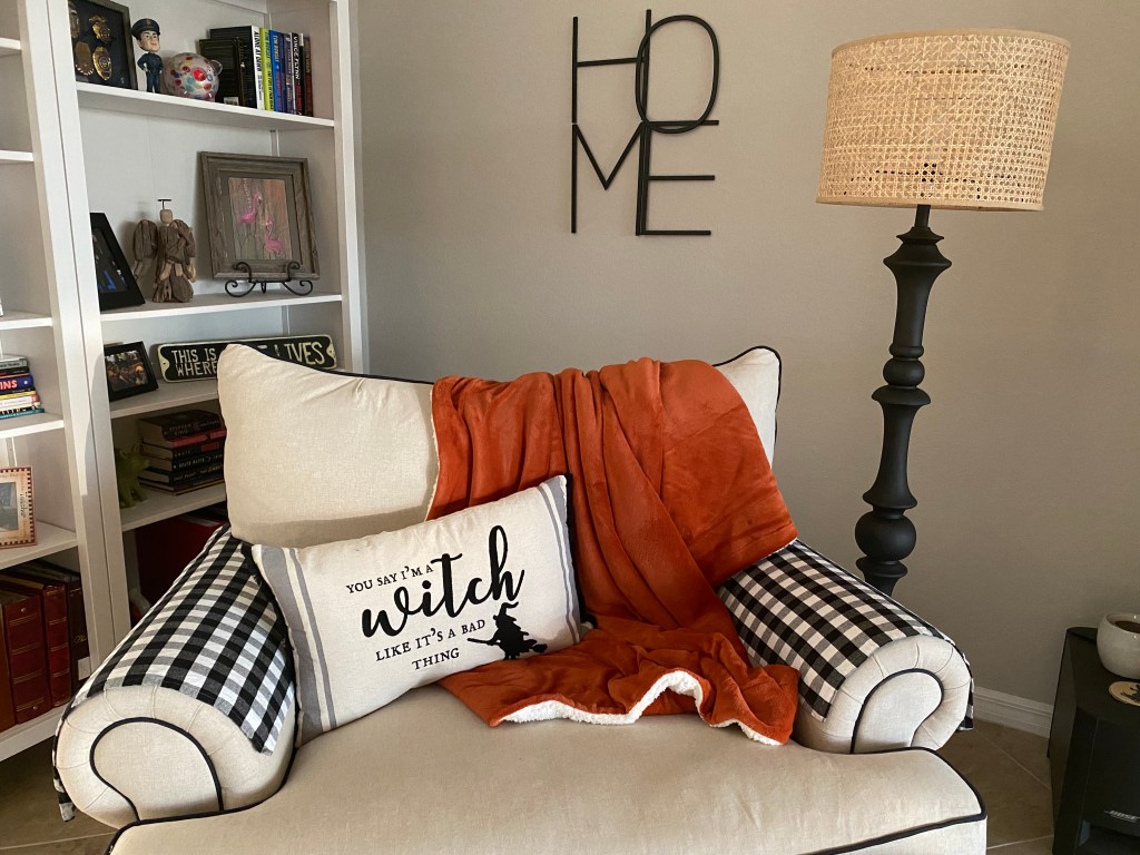 A cozy living room corner featuring an armchair with a black and white checkered armrest, an orange throw blanket draped over it, and a pillow that reads 'You say I'm a witch like it's a bad thing'. A woven lamp and a bookshelf filled with books and decorative items complete the scene.