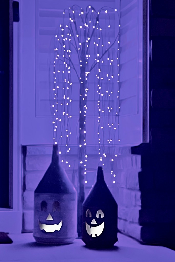 A cozy front porch featuring two whimsical lanterns made from old oil cans, with glowing pumpkin faces and a whimsical purple haunted tree adorned with fairy lights in the background.