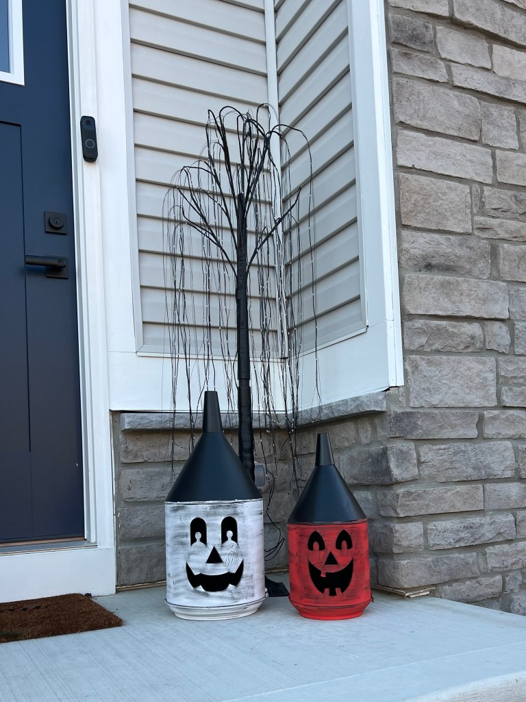 Two whimsical Halloween decorations, featuring black and white painted lanterns shaped like jack-o'-lanterns, positioned beside a black haunted tree, set against a stone wall and a front door.