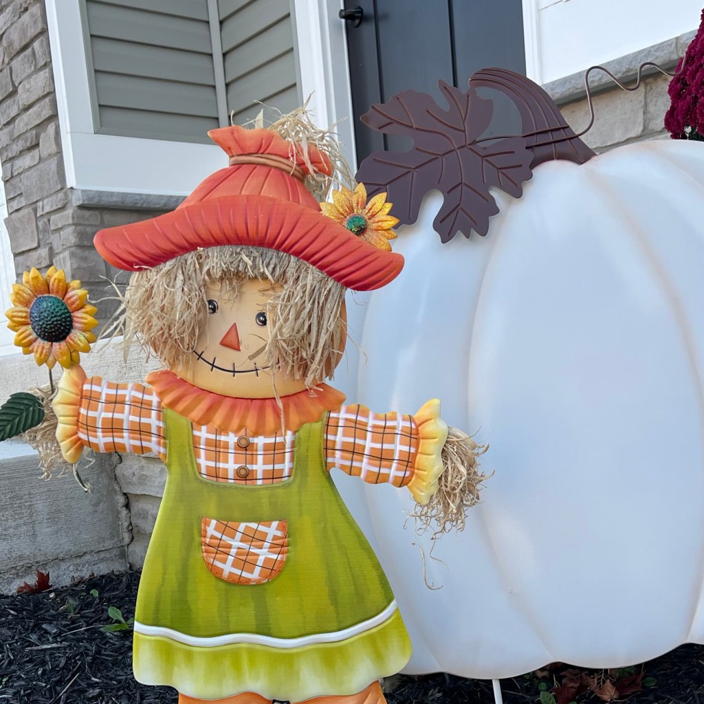 A cheerful scarecrow with a sunflower stands next to a large white pumpkin, decorated with vibrant colors in an autumn-themed front porch scene.