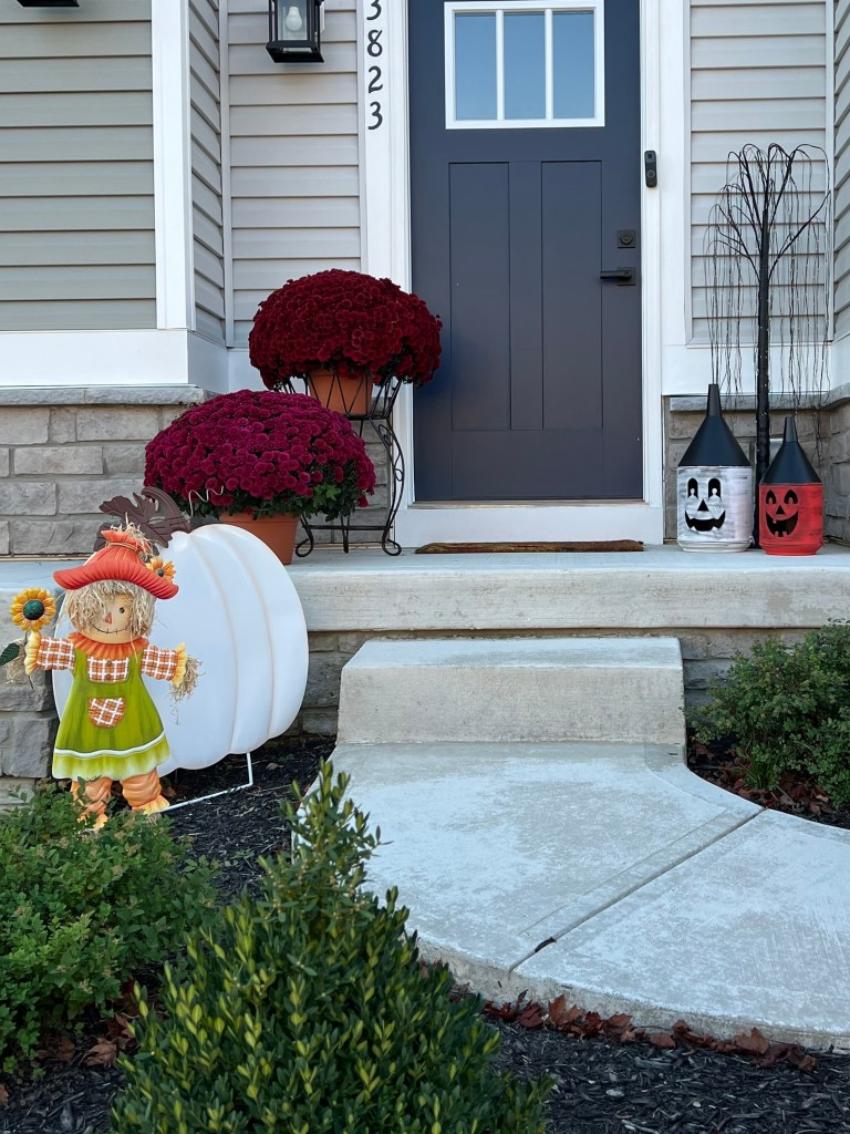 A front porch decorated for autumn, featuring vibrant purple mums in planters, a cheerful scarecrow holding a sunflower, a large white pumpkin, and whimsical lanterns with Halloween designs.