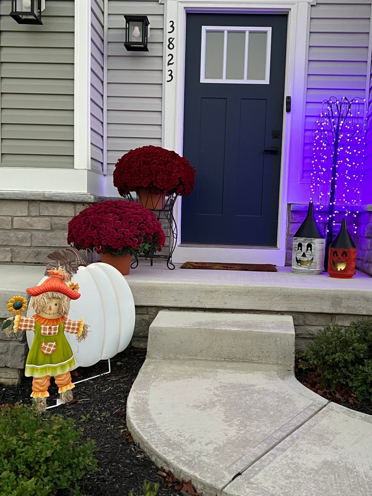 A cozy front porch decorated for autumn, featuring vibrant red mums, a cheerful scarecrow, a large white pumpkin, and glowing lanterns with Halloween designs, set against a blue door.