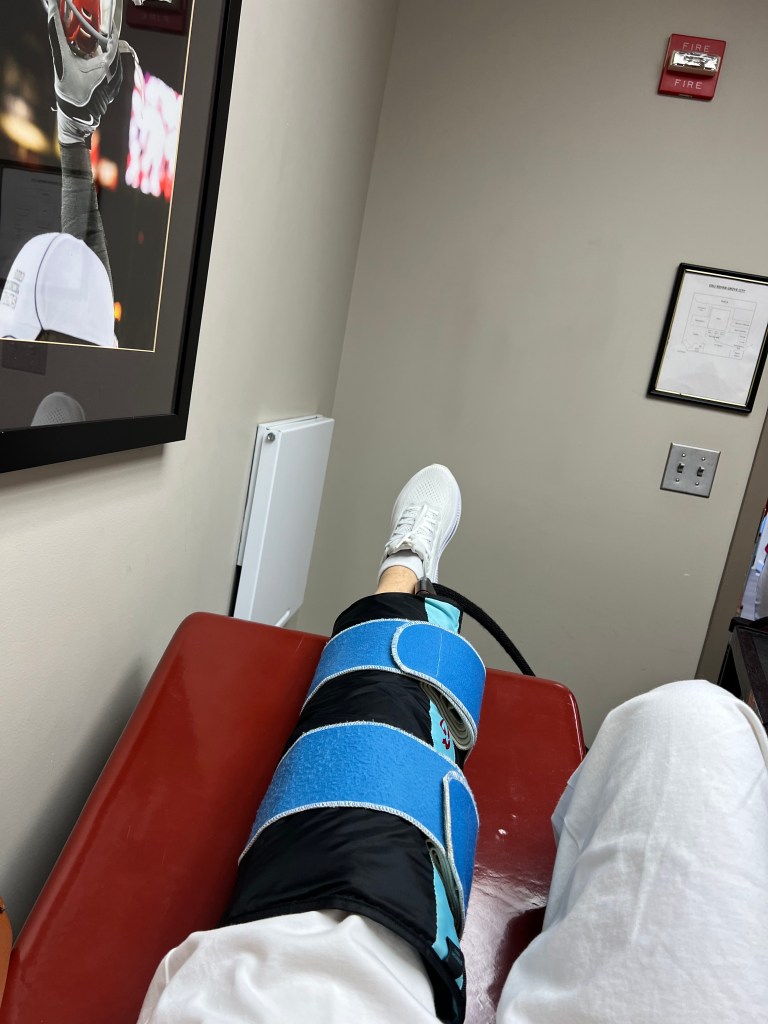 A leg in a knee brace resting on a treatment table in a medical setting, with a framed sports photo in the background.